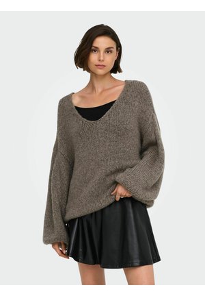 Femme aux cheveux courts bruns portant un pull en laine gris ample par-dessus un haut noir et une jupe en cuir courte noire, debout devant un fond clair.