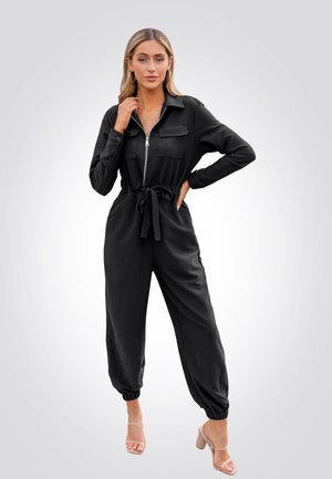 Combinaison noire avec fermeture éclair sur le devant, poches poitrine boutonnées, taille ceinturée et poignets fuselés. Fabriquée dans un tissu lisse avec une coupe décontractée.