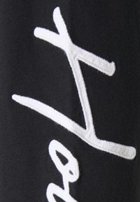 Svart tyg med en framträdande vit broderad textdesign. Texturen är mjuk och bokstäverna har en upphöjd, plysch känsla.