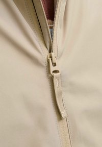 Beige jakke med en glatt tekstur, med en halvglidelås og en kontrasterende indre stoff. Glidelåstrekket er over en søm.