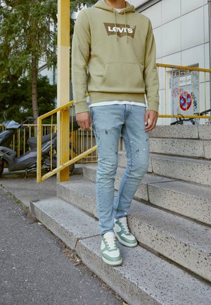 Osoba ubrana w jasnozieloną bluzę z kapturem Levi's, przetarte jasnoniebieskie dżinsy i białozielone buty sportowe stojąca na zewnętrznych betonowych schodach.