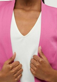 Růžový sako přes bílé tričko s výstřihem ve tvaru V, hladká tkanina, přesný střih, s rukama umístěnými na límci, zvýrazňující dekolt.