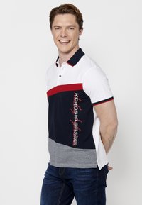 Cămașă polo cu mâneci scurte, disponibilă în alb, bleumarin, roșu și gri, dotată cu nasturi. Prezintă un design de logo îndrăzneț și blocuri de culoare contrastante.