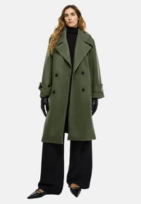 Grüner Wollmischung-Doppelreiher-Mantel mit großen Spitzen, schwarzen Knöpfen und hochgerollten Ärmeln, getragen über einem schwarzen Rollkragenpullover und weit geschnittenen Hosen.