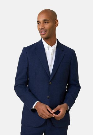 Mann trägt einen dunkelblauen Anzug und ein weißes Hemd, schaut leicht nach links mit locker verschränkten Händen.