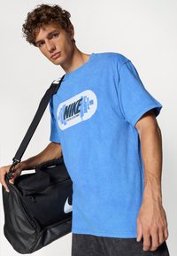 Blå bomulds T-shirt med grafisk tryk, der viser ordet "NIKE" og et vægt-design. Sort træningstaske med et hvidt Nike-logo.