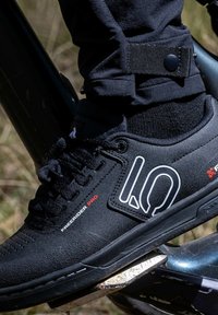 Scarpa da ciclismo nera con una tomaia strutturata, caratterizzata da un logo bianco e un accento rosso. La scarpa è fissata con i lacci e posizionata su un pedale.