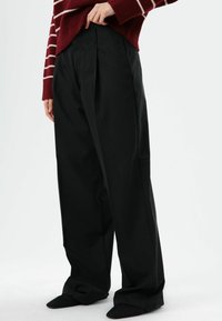 Zwarte geplooide broek met een losse pasvorm, voorzien van een gladde textuur en een hoge taille. Gecombineerd met donkere schoenen met een puntige neus.