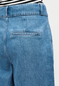 Οπίσθια όψη ατόμου που φοράει ανοιχτό γαλάζιο τζιν με μια σχισμή σε πίσω τσέπη και μια μαύρο-άσπρη ριγέ πουκάμισο που είναι μέσα στο παντελόνι.