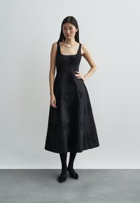 Czarna sukienka bez rękawów z dopasowanym gorsetem i pełną spódnicą, zdobiona misternymi, koralikowymi wzorami kwiatowymi. Noszona z czarnymi rajstopami i butami.