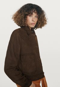 Bruine suèdejas met een kraag, twee grote zakken aan de voorkant en ronde knopen. Heeft een losse pasvorm en een gladde textuur.