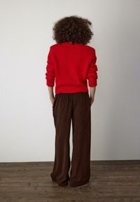 Czerwony sweter z dzianiny o luźnym kroju i ściągaczach przy rękawach, zestawiony z brązowymi szerokimi spodniami welurowymi z zakładkami i kieszeniami po bokach.