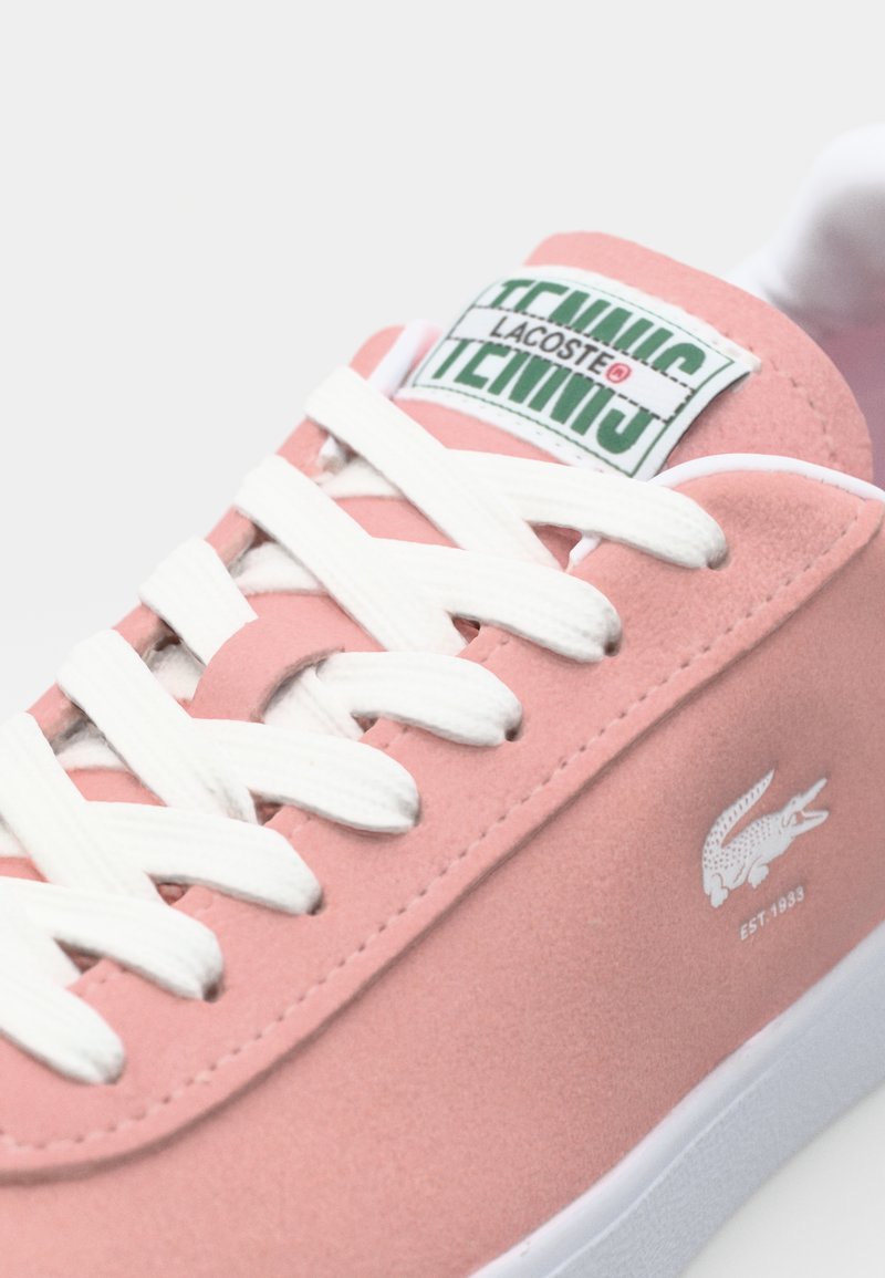 Zapatillas de ante rosa claro con cordones blancos, que presentan una etiqueta verde y blanca de "TENNIS" en la lengüeta y un pequeño logo de cocodrilo blanco en el lateral.