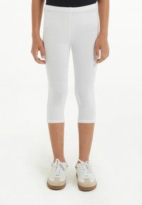 Leggings capri blancs en tissu extensible, design ajusté, longueur mi-mollet ; associés à des baskets blanches et beiges avec lacets.