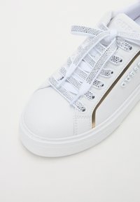 Zapatilla de cuero blanca con punta redondeada, acentuada con una franja dorada, con cordones adornados con cristales y suela plana de goma.