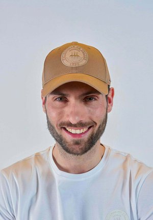 Homme souriant avec une courte barbe foncée portant une casquette de baseball marron avec un logo rond et un t-shirt blanc sur un fond clair uni.