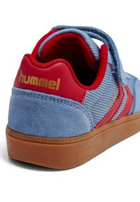 Niebieski zamszowo-materiałowy tenisówka z czerwonymi akcentami, wyposażona w wyściełany kołnierz, pasek na rzep oraz teksturowaną gumową podeszwę w kolorze piaskowym. Złote logo.