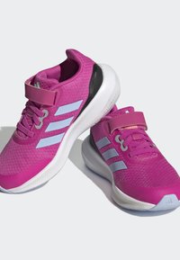 Zapatillas deportivas rosas con parte superior de tela texturizada, entresuela blanca, acentos negros y tres rayas azul claro a cada lado. Incluye una correa ajustable.