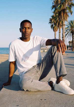 Muž v bílém tričku, šedých džínách a bílých teniskách sedí na chodníku poblíž pláže s palmami pod jasně modrou oblohou.