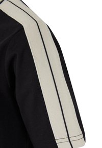 Svart t-skjorte med en strukturert beige stripe langs skulderen, akkompagnert av en tynn svart linje. Stoffet ser mykt og pustende ut.