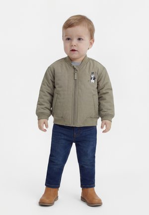 Småbarn iført en beige bomberjakke med tegneserie-patch, blå jeans og beige støvler, stående mod en hvid baggrund.