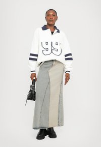 Woman wearing white rugby-style top with number 99, two-tone patchwork denim maxi skirt, black boots, holding small black handbag.