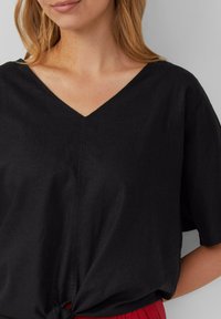 Mustan V-kaeluse avec lühikesed varrukad ja sõlmega detail hemline'il. Sile, kergesti langev kangas peene tekstuuriga.