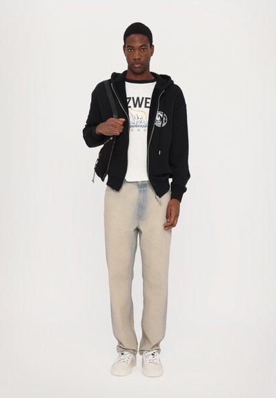 Black zip-up hoodie, white graphic t-shirt, light gray jeans, and white sneakers. The model is holding a small black bag.