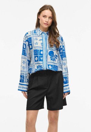 Vrouw draagt een blauw-wit patroon lange mouwen shirt met zwarte shorts met hoge taille, staand tegen een effen witte achtergrond.