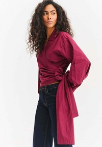 Bordowa koszula kopertowa z długimi rękawami, z wiązaniem w talii i gładką fakturą, zestawiona z ciemnymi jeansami.