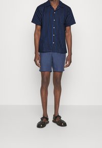 Camicia a maniche corte blu navy con righe verticali blu, abbinata a pantaloni corti blu scuro e sandali neri intrecciati con dettagli metallici.