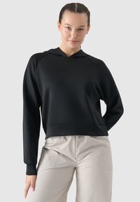 Felpa nera con cappuccio, maniche lunghe a raglan, texture liscia e taglio corto. Indossata con pantaloni chiari e larghi.
