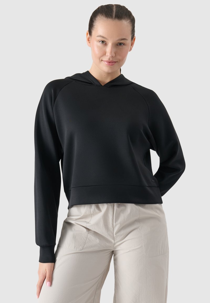 Felpa nera con cappuccio, maniche lunghe a raglan, texture liscia e taglio corto. Indossata con pantaloni chiari e larghi.
