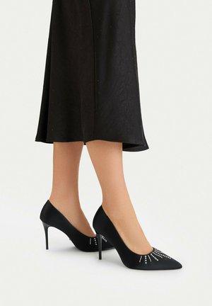 Mujer con zapatos de tacón alto negros con tachuelas decorativas plateadas, combinados con un vestido negro de longitud media.