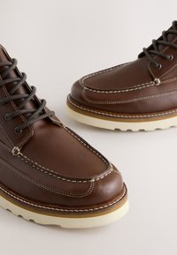 Ruskeat nahkakengät harmailla nauhoilla, joissa on mokkakenkien kärkipitsin muotoilu, valkoinen ompelu ja paksu kermanvärinen pohja, jossa on ruskea yksityiskohta.