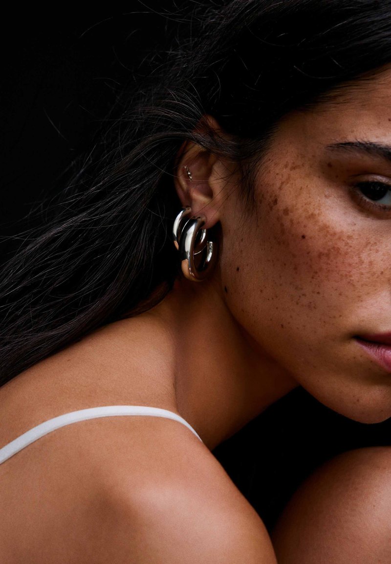 Gros plan d'une femme aux cheveux foncés, avec des taches de rousseur, des boucles d'oreilles argentées en forme de créoles et une bretelle blanche sur son épaule sur fond noir.