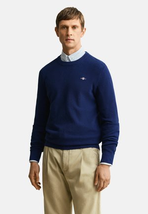 Man draagt een marineblauwe trui over een lichtblauw overhemd met knopen en beige broek, staand tegen een effen lichte achtergrond.