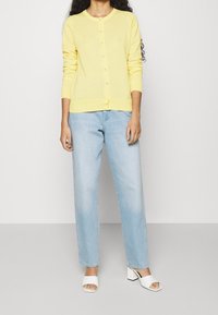 Gul stickad cardigan med rund halsringning och knappar framtill, kombinerad med ljusblå raka jeans och vita klacksandaler.