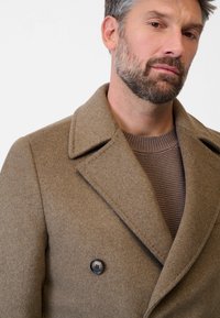 Brauner Woll-Peacoat mit doppelter Knopfreihe, breitem Kragen und schwarzem Knopfdetail, über einem braunen strukturierten Strickpullover getragen.