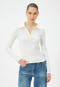 Maglietta bianca a maniche lunghe con collo e bottoni. Presenta una texture a coste e un design aderente. Indossata con jeans blu.