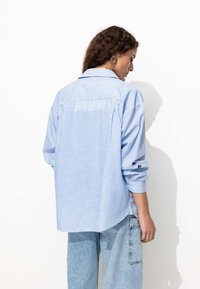 Chemise à rayures bleu clair à manches longues avec un col, coupe décontractée, manches retroussées et un empiècement dans le dos. Associée à un pantalon en denim clair.