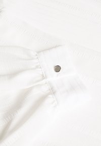 Tessuto bianco texture con strisce verticali, con polsino e dettaglio della chiusura a pressione argentata.