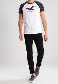 Vit bomullströja med grå raglanärmar och en vinröd "Hollister"-logotyp. Bärs med svarta jeans och grå sneakers.