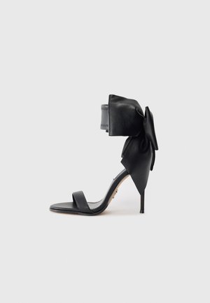 Black high-heel sandal made of leather, featuring a wide ankle strap with a large bow and a slender heel. Minimalist design.