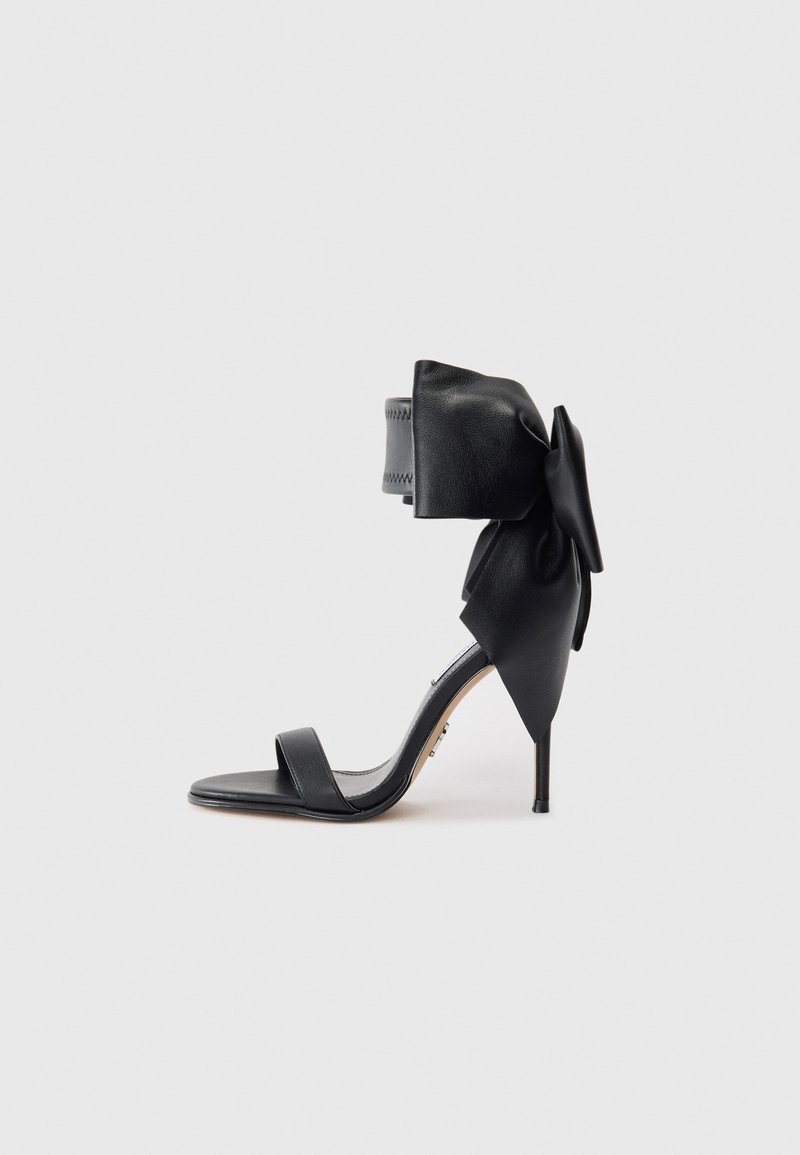 Black high-heel sandal made of leather, featuring a wide ankle strap with a large bow and a slender heel. Minimalist design.