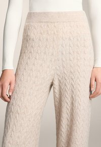 Nohavice so vzorom pleteniny v svetlobéžovej farbe, s rebrovaným pásom a mäkkým, textúrovaným vzhľadom. Materiál pôsobí teplo a pohodlne.
