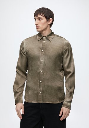 Jonge man met kort bruin haar die een lange mouwen olijfgroene satijnen blouse met knopen en zwarte broek draagt, kijkt zijwaarts tegen een witte achtergrond.