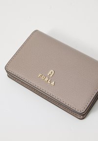 Lederen portemonnee in licht taupe met een gegranuleerde textuur. Heeft een gouden "FURLA" logo, contrasterende stiksel en een compacte rechthoekige vorm.