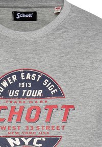 Camiseta de algodón gris con cuello redondo, que presenta un estampado gráfico en rojo y azul marino. El texto incluye "Schott" y "Lower East Side U.S. Tour."