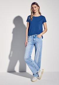 Blå t-shirt med kort ärm, ljus tvättade vida jeans och vita sneakers. Slätt tyg, avslappnad passform och subtil design utan synliga mönster.